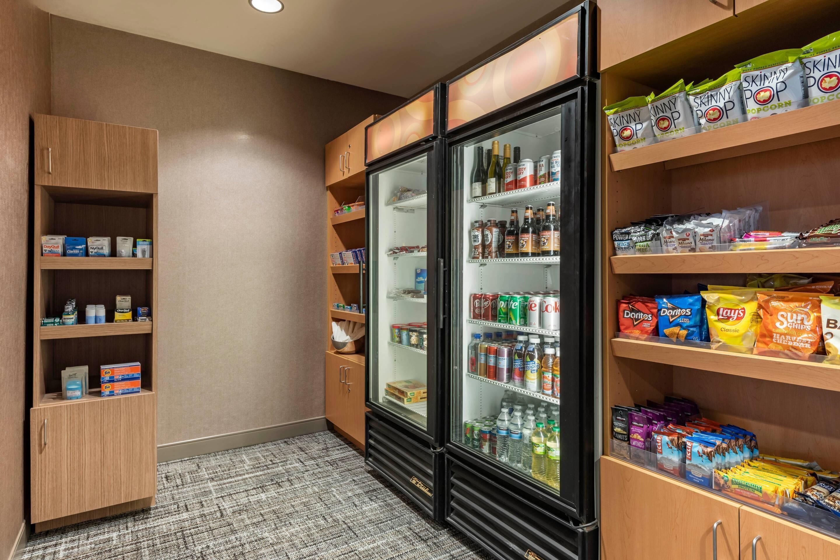 Une chambre d’hôtel équipée d’un mini-bar, d’un réfrigérateur rempli de boissons, ainsi que d’étagères contenant des en-cas et des articles de toilette.