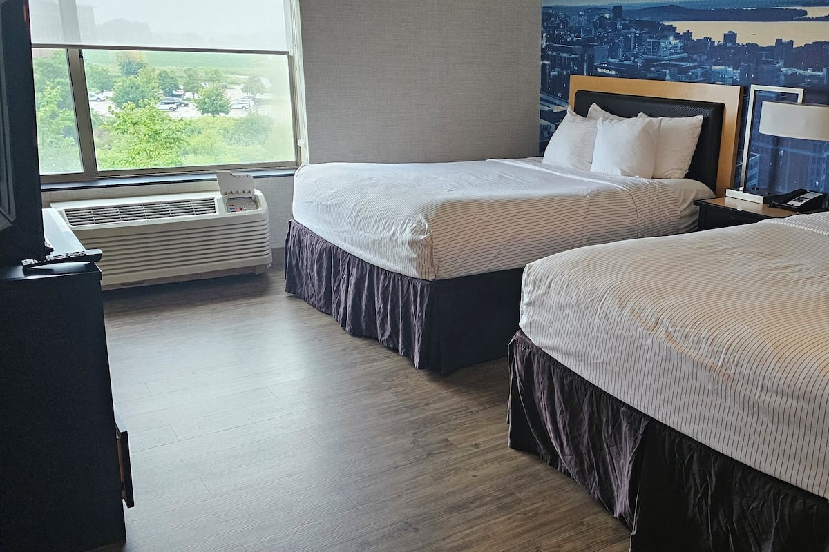 Habitación de hotel con dos camas, un ventanal grande, un mural con vista a la ciudad y un escritorio con televisor.