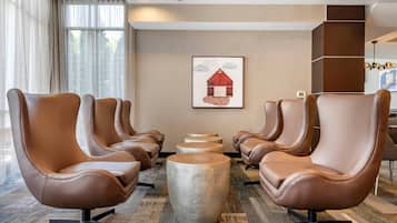 Una zona de estar con cuatro sillones de cuero marrón y tres mesas redondas, un cuadro enmarcado en la pared y una ventana con cortinas translúcidas.