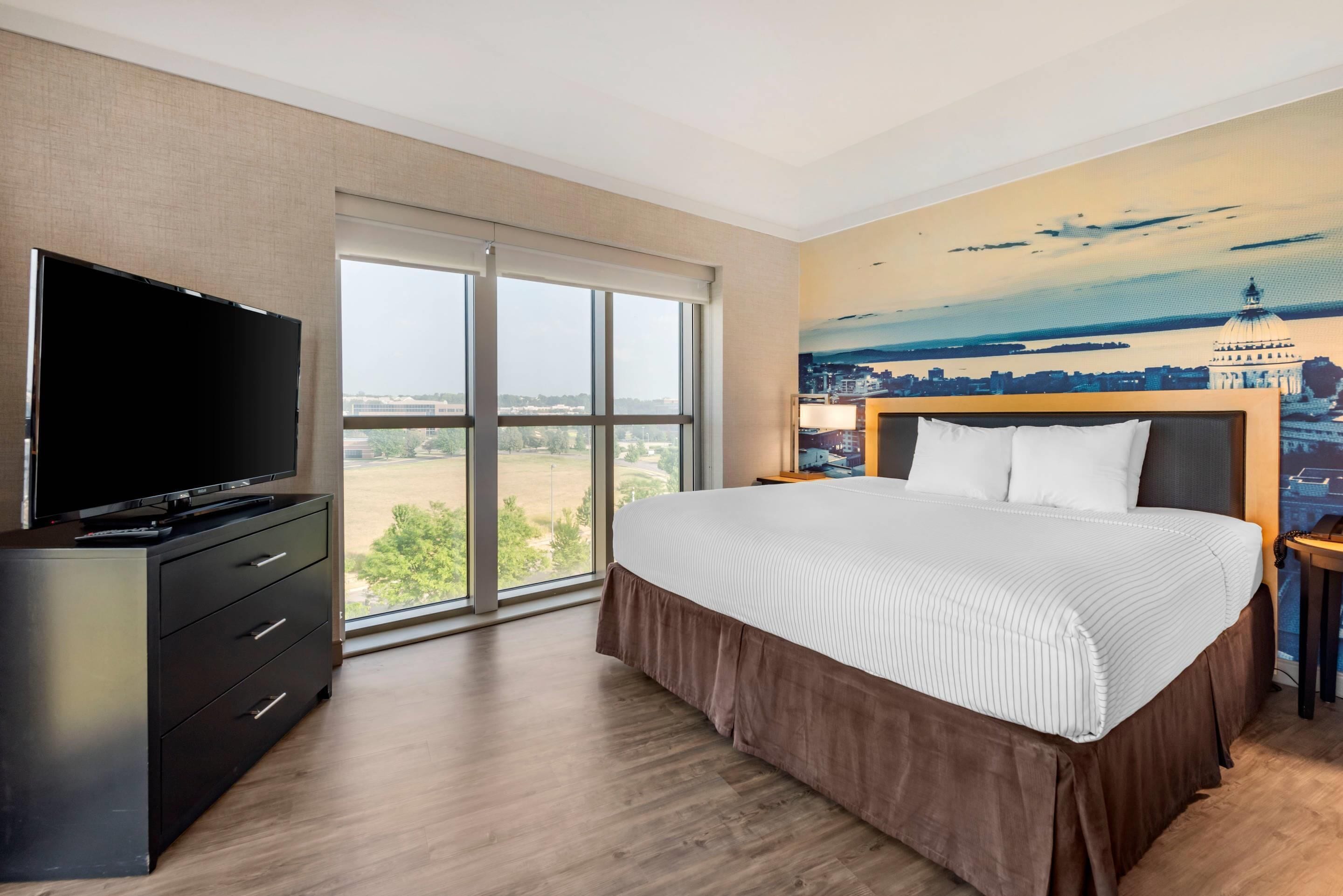 Habitación de hotel con una cama grande, un televisor en una cómoda, un mural con un paisaje urbano y vistas por la ventana.