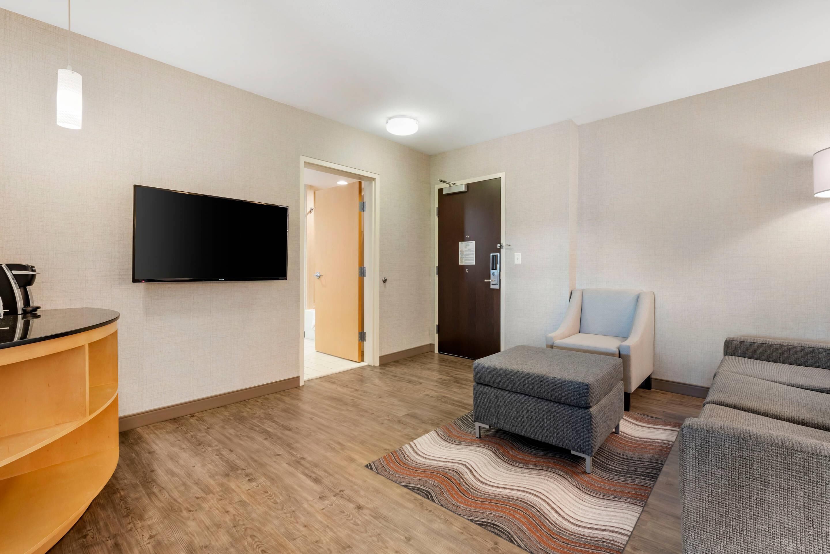 A modern hotel room with a flat-screen TV mounted on the wall, a wooden floor, a patterned rug, a grey ottoman, and a sofa.
