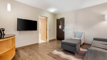 A modern hotel room with a flat-screen TV mounted on the wall, a wooden floor, a patterned rug, a grey ottoman, and a sofa.