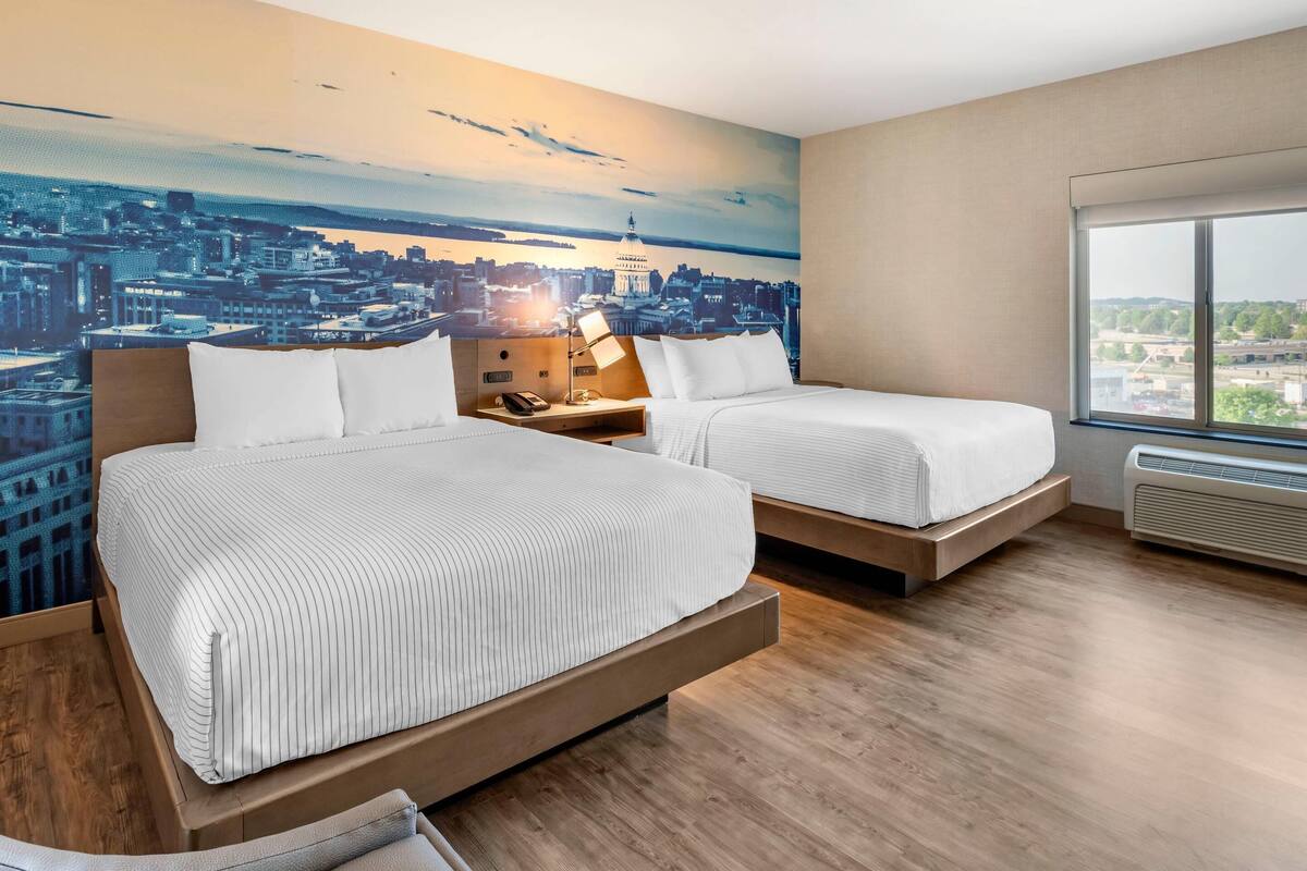 Habitación de hotel con dos camas, un mural con vista a la ciudad en la pared, un ventanal grande y un diseño moderno.