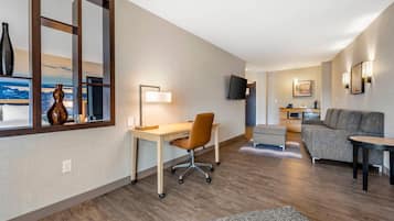 A modern hotel room with a wooden desk, a comfortable sofa, a flat-screen TV, and a city view through a large window.