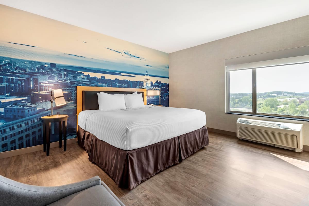 Habitación de hotel con una cama grande, una mesita de noche, una lámpara, una ventana con vistas a un paisaje urbano y un equipo de aire acondicionado.