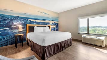 A hotel room with a large bed, a nightstand, a lamp, a window with a view of a cityscape, and an air conditioning unit.