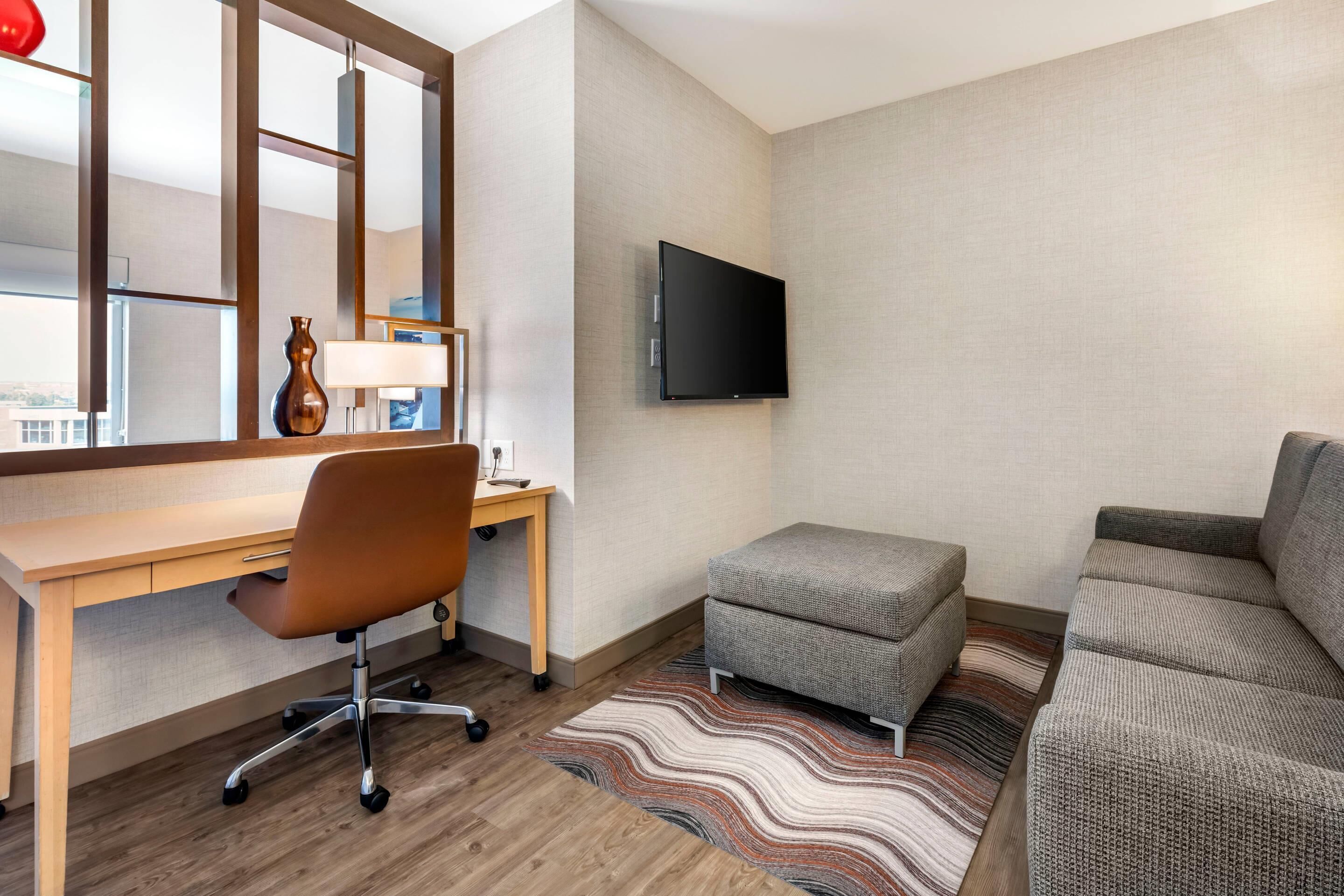 Una habitación de hotel con un escritorio de madera, una silla de oficina color café, un sofá gris, un televisor de pantalla plana montado en la pared y un tapete a rayas en el suelo.