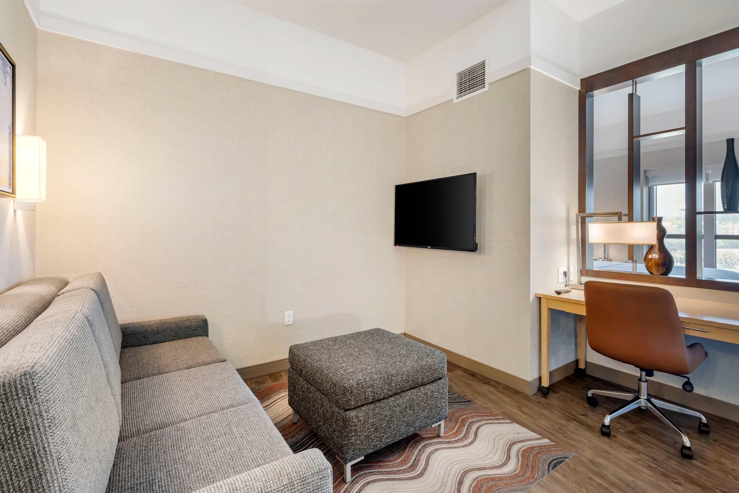 A modern hotel room with a grey sofa, a small grey ottoman, a wooden desk with a chair, and a flat-screen TV mounted on the wall.