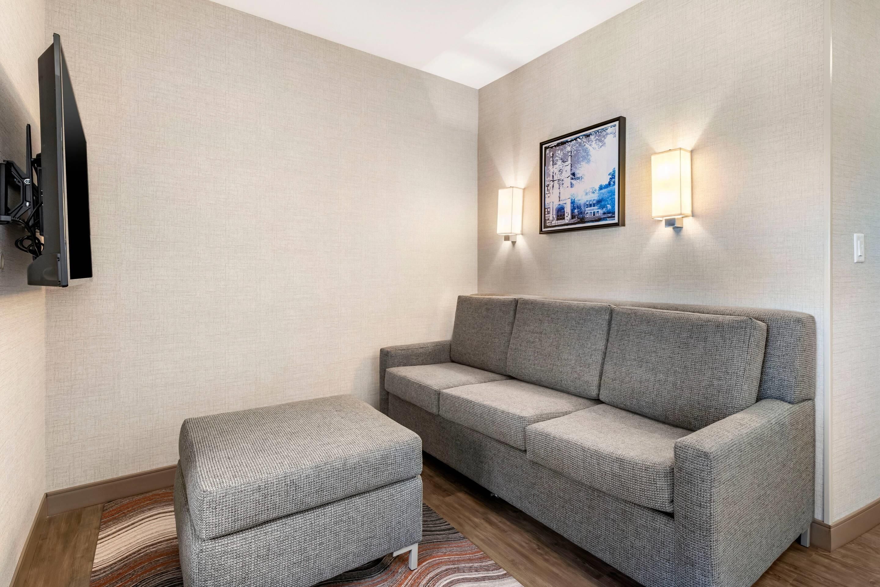 A modern living room with a grey sofa, a matching ottoman, a flat-screen TV mounted on the wall, and two wall-mounted lamps.