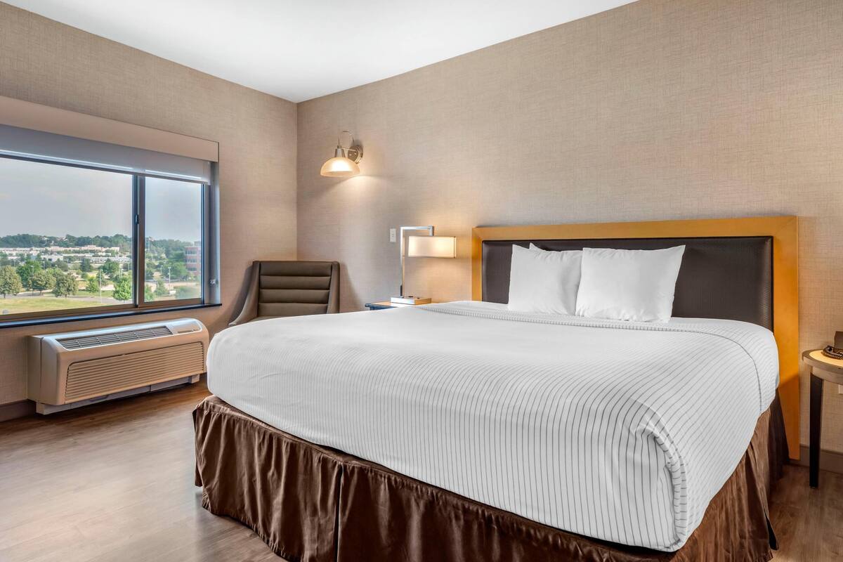 Habitación de hotel con una cama grande, una silla, una mesita de noche, una lámpara y vistas a la ciudad a través de un ventanal amplio.