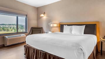 Habitación de hotel con una cama grande, una silla, una mesita de noche, una lámpara y vistas a la ciudad a través de un ventanal amplio.