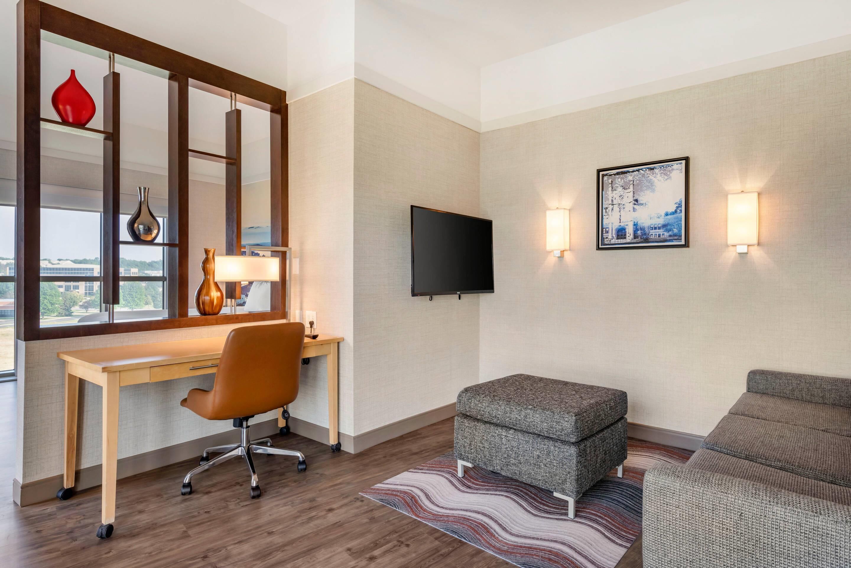 Quarto de hotel com escrivaninha de madeira, cadeira moderna, sofá cinza, tapete estampado, televisão de tela plana e luminárias fixadas na parede.