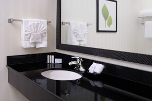 A modern bathroom with a black countertop, a white sink, and a large mirror. There are two white towels hanging on towel bars, and a framed picture of leaves on the wall. - Fairfield Inn & Suites by Marriott Dallas Plano/The Colony (The Colony)