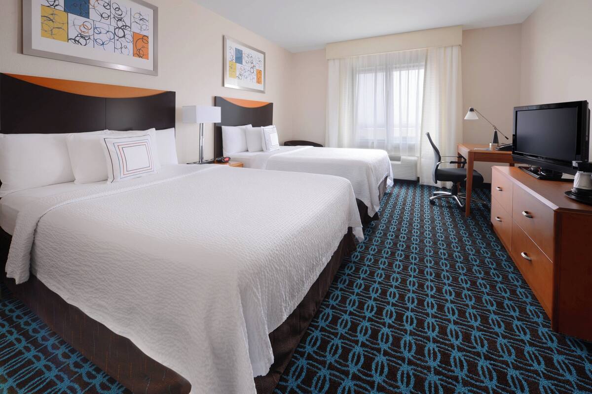 A hotel room with two beds, a desk with a television, and a patterned carpet.