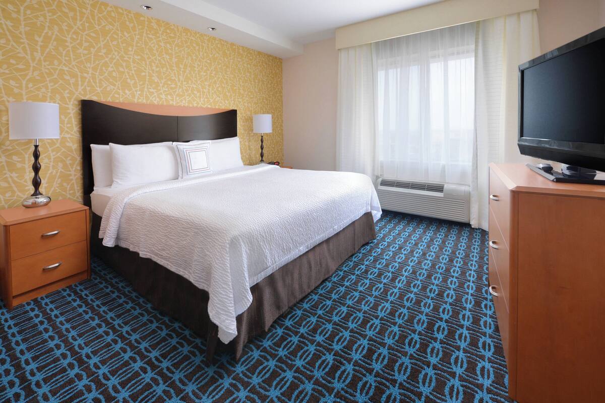 A hotel room with a large bed, bedside lamps, a flat-screen TV, a dresser, and a window with curtains.