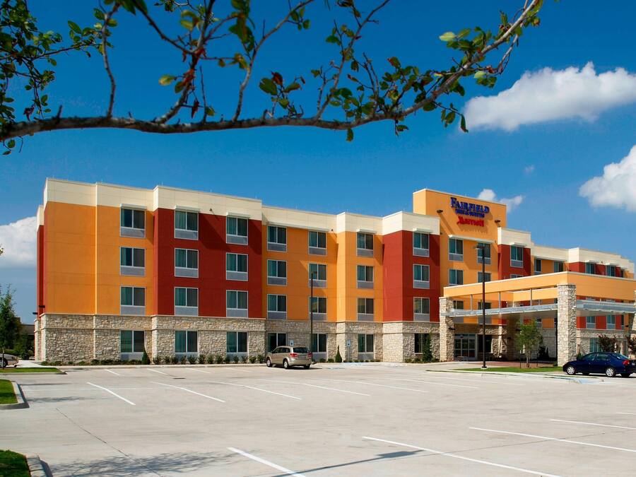 A multi-story hotel with a modern design, featuring a mix of orange and beige colors, a parking lot, and a clear blue sky.