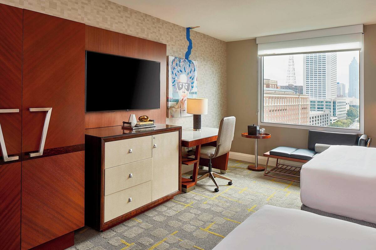 Ein Hotelzimmer mit einem großen Bett, einem Schreibtisch mit Stuhl, einem an der Wand befestigten Fernseher und einem Fenster mit Blick auf die Stadt.