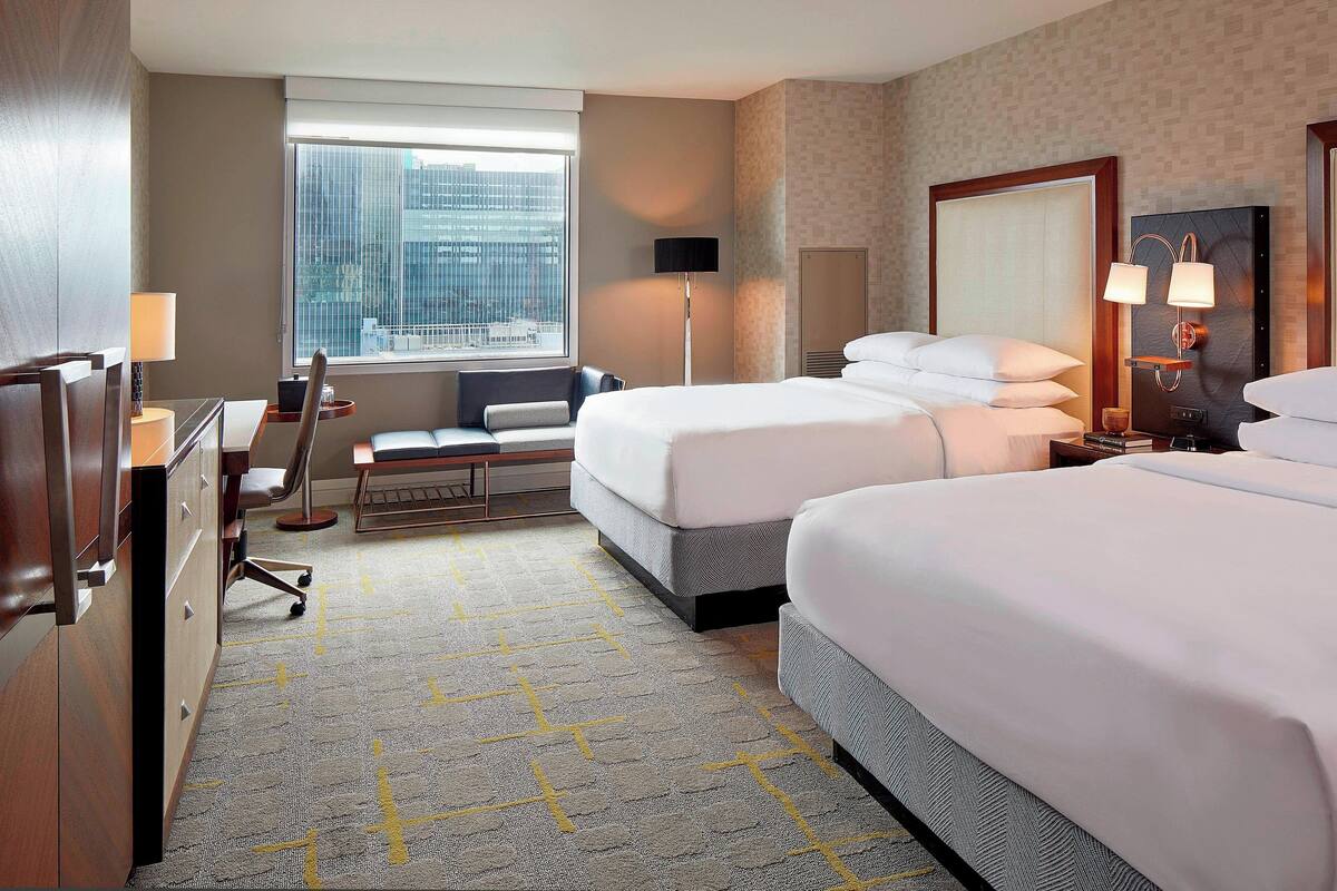 Ein Hotelzimmer mit zwei Betten, einem Schreibtisch, einem Stuhl, einer Lampe und Blick auf die Stadt durch das Fenster.