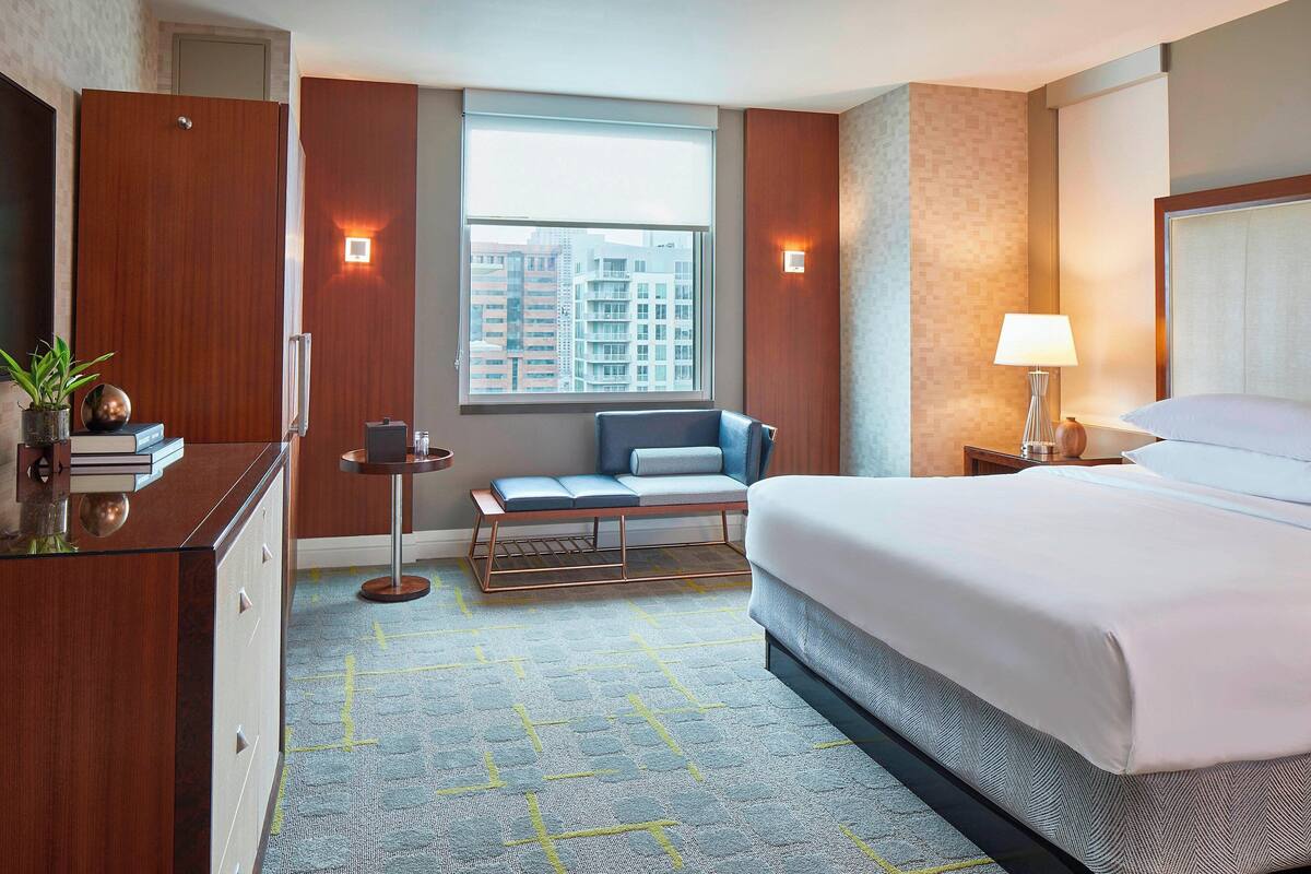 Ein Hotelzimmer mit einem großen Bett, einem Schreibtisch, einem Stuhl, einem kleinen Tisch und einem Fenster mit Blick auf Gebäude.
