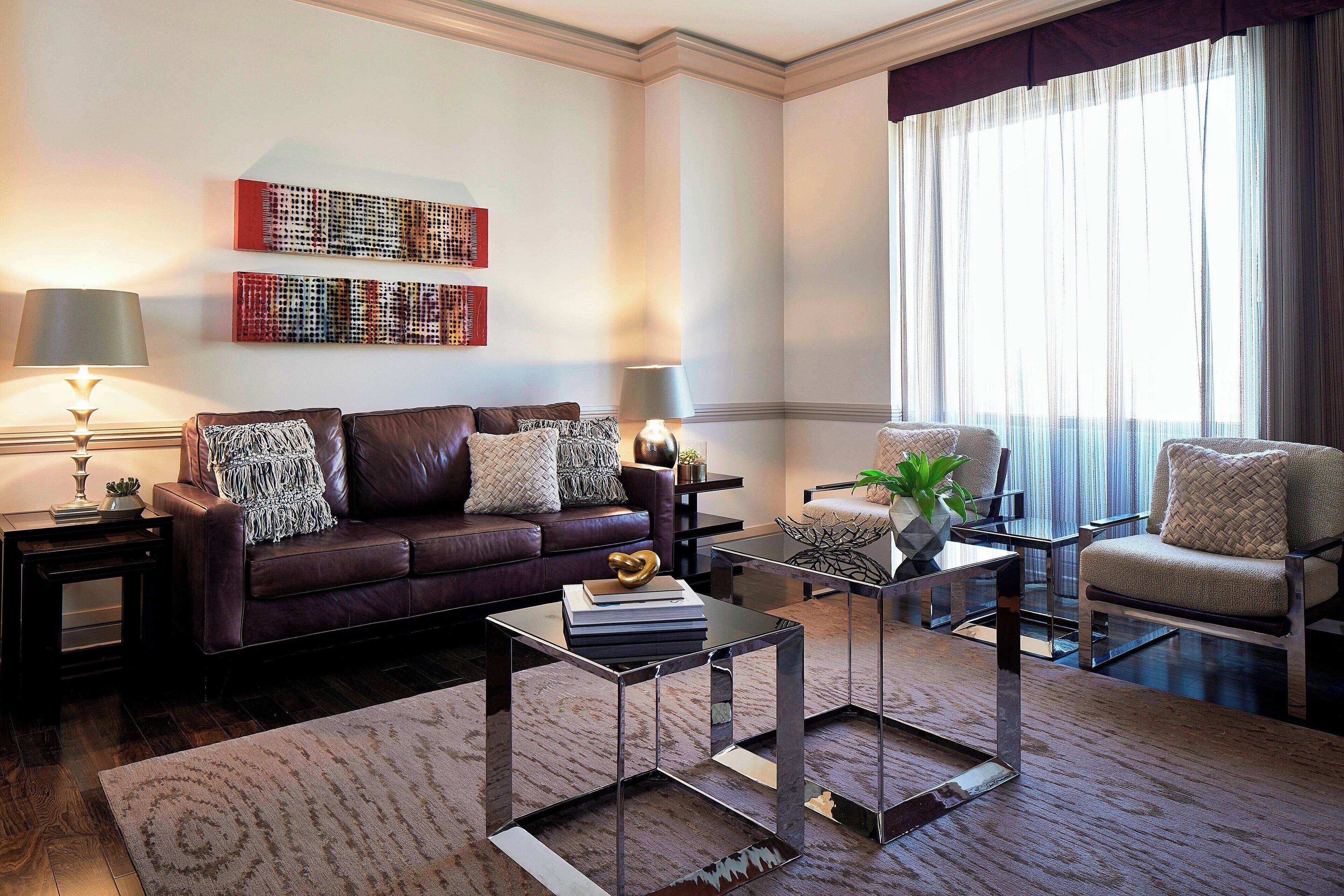 A living room with a dark brown leather sofa, two armchairs, a coffee table, and a rug. There are two lamps on side tables, and artwork on the wall.