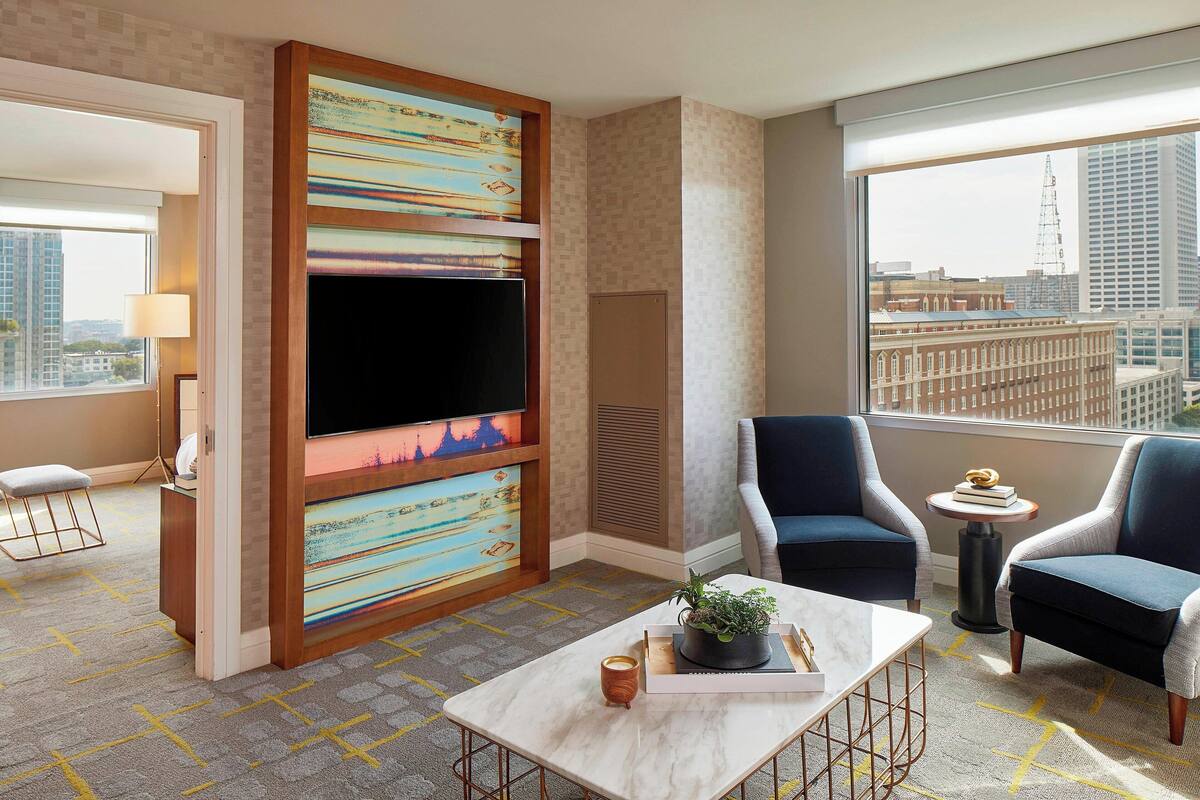 Ein modernes Hotelzimmer mit großem Fernseher, zwei Sesseln, einem kleinen Tisch mit Pflanze und Blick auf die Stadt durch das Fenster.