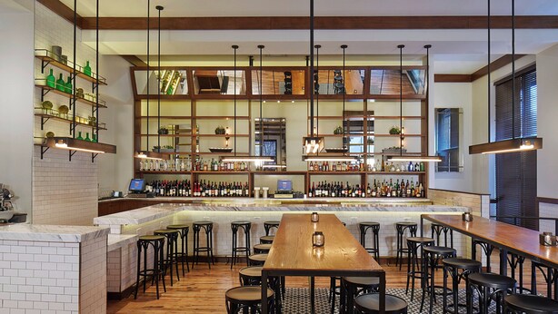 A bar area with a long wooden table, multiple bar stools, and shelves stocked with bottles. - Renaissance Atlanta Midtown Hotel (Atlanta)