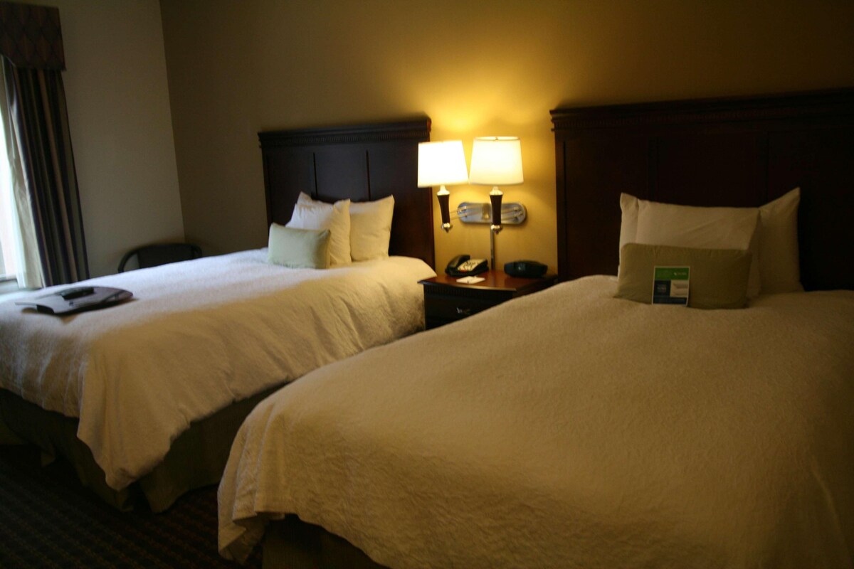 A hotel room with two beds, a nightstand with a lamp, and a chair.