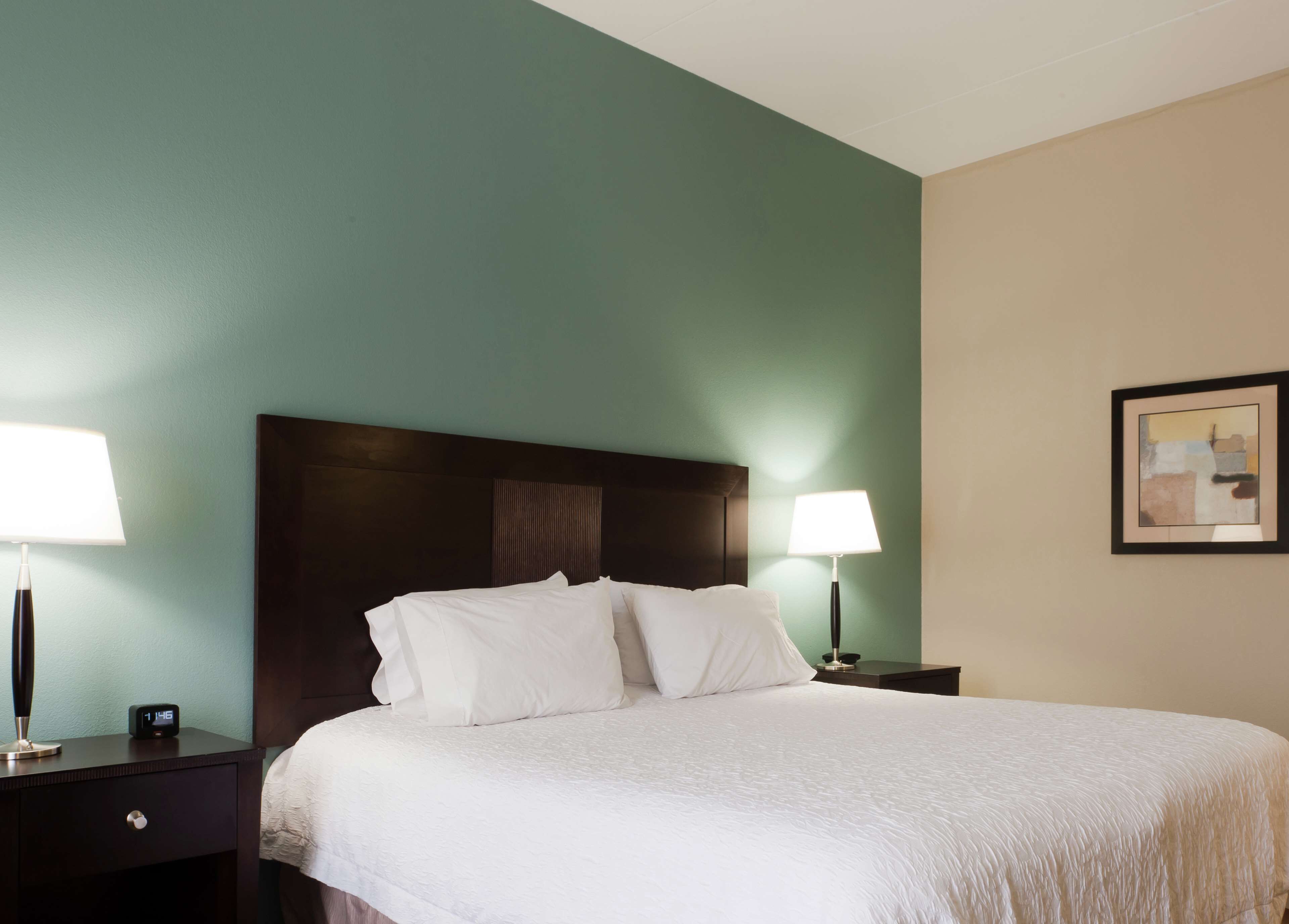 A hotel room with a bed, two bedside tables, a nightstand, a wall clock, and a framed picture.