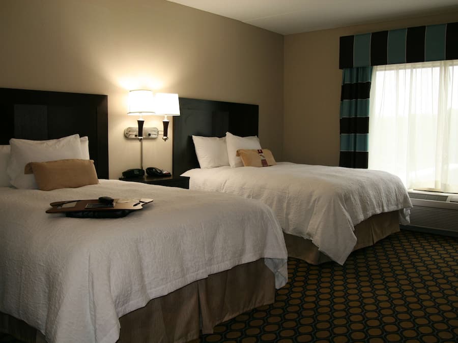 A hotel room with two beds, a nightstand with a lamp, a telephone, and a window with curtains.