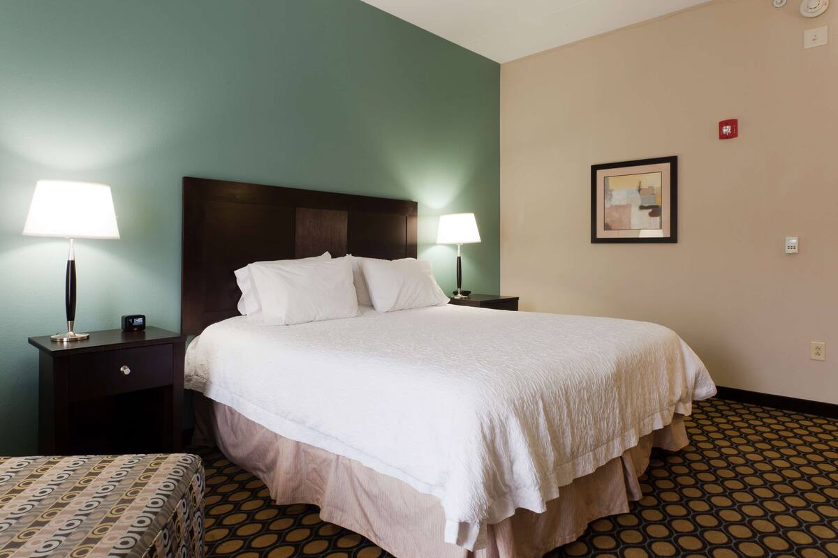 A hotel room with a large bed, two bedside tables with lamps, a headboard, a framed picture on the wall, and a patterned floor.