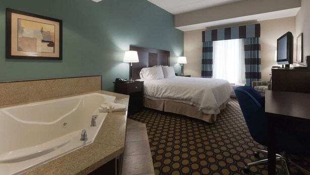 A hotel room with a large bed, a desk, a chair, a desk lamp, a TV, a bathroom with a bathtub, and a framed picture on the wall. - Hampton Inn North Brunswick / New Brunswick (North Brunswick)