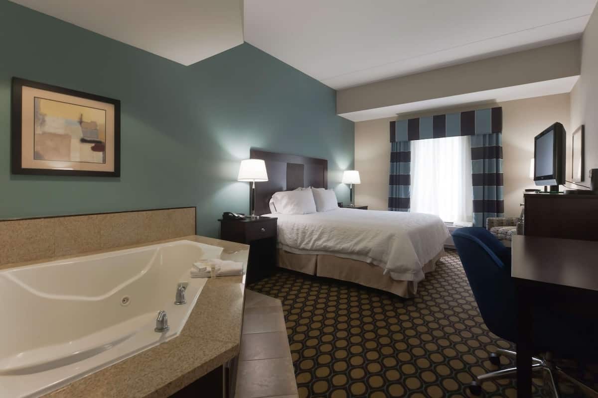 A hotel room with a large bed, a desk, a chair, a desk lamp, a TV, a bathroom with a bathtub, and a framed picture on the wall.