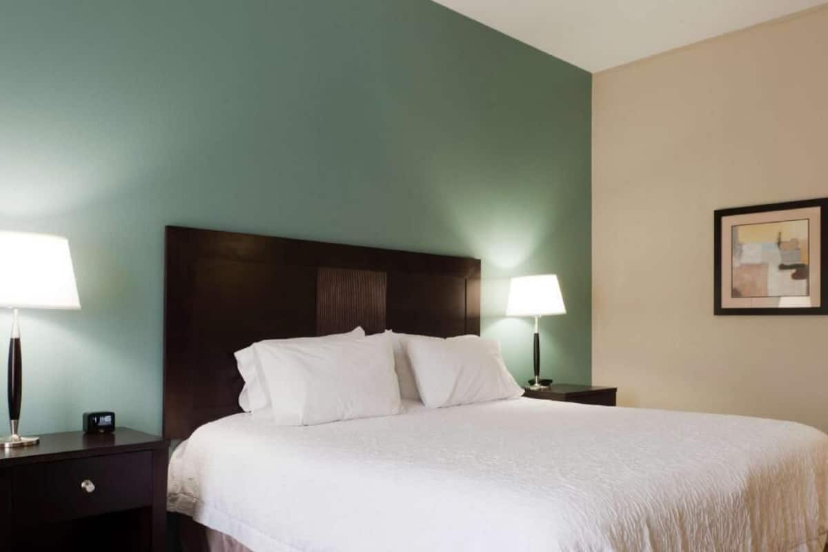 A hotel room with a large bed, two bedside tables with lamps, a framed picture on the wall, and a dark headboard.