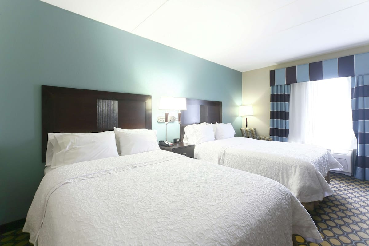 A hotel room with two beds, a nightstand with a lamp, a telephone, and a window with curtains.