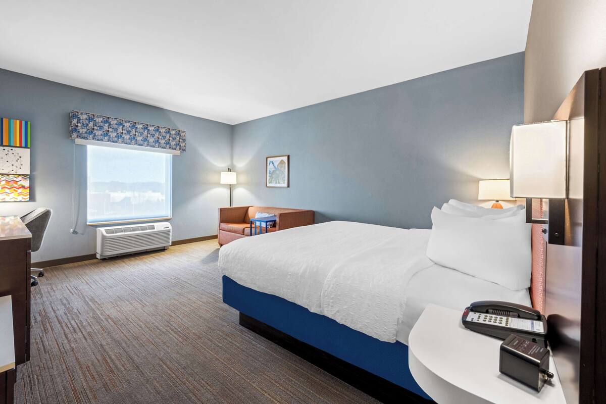 A hotel room with a large bed, a desk, a chair, a window with a view, and a wall-mounted telephone.