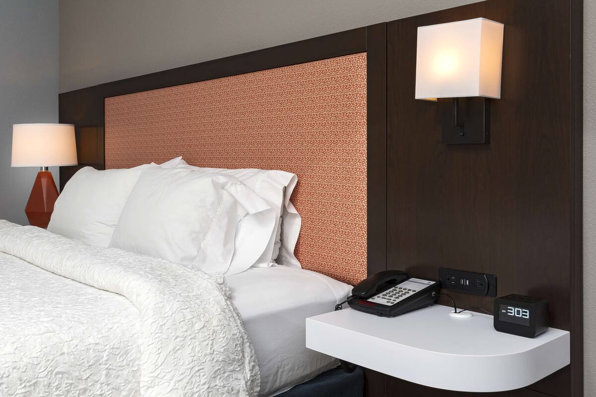 A hotel room with a neatly made bed, a bedside table with a phone and a digital clock, and two wall-mounted lamps.