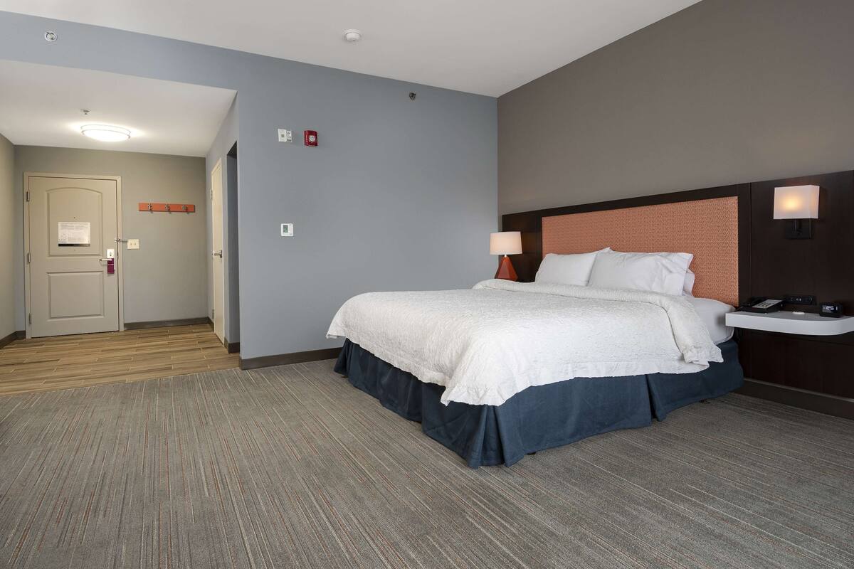 A hotel room with a large bed, a bedside table with a lamp, a wall-mounted phone, and a small desk with a chair.