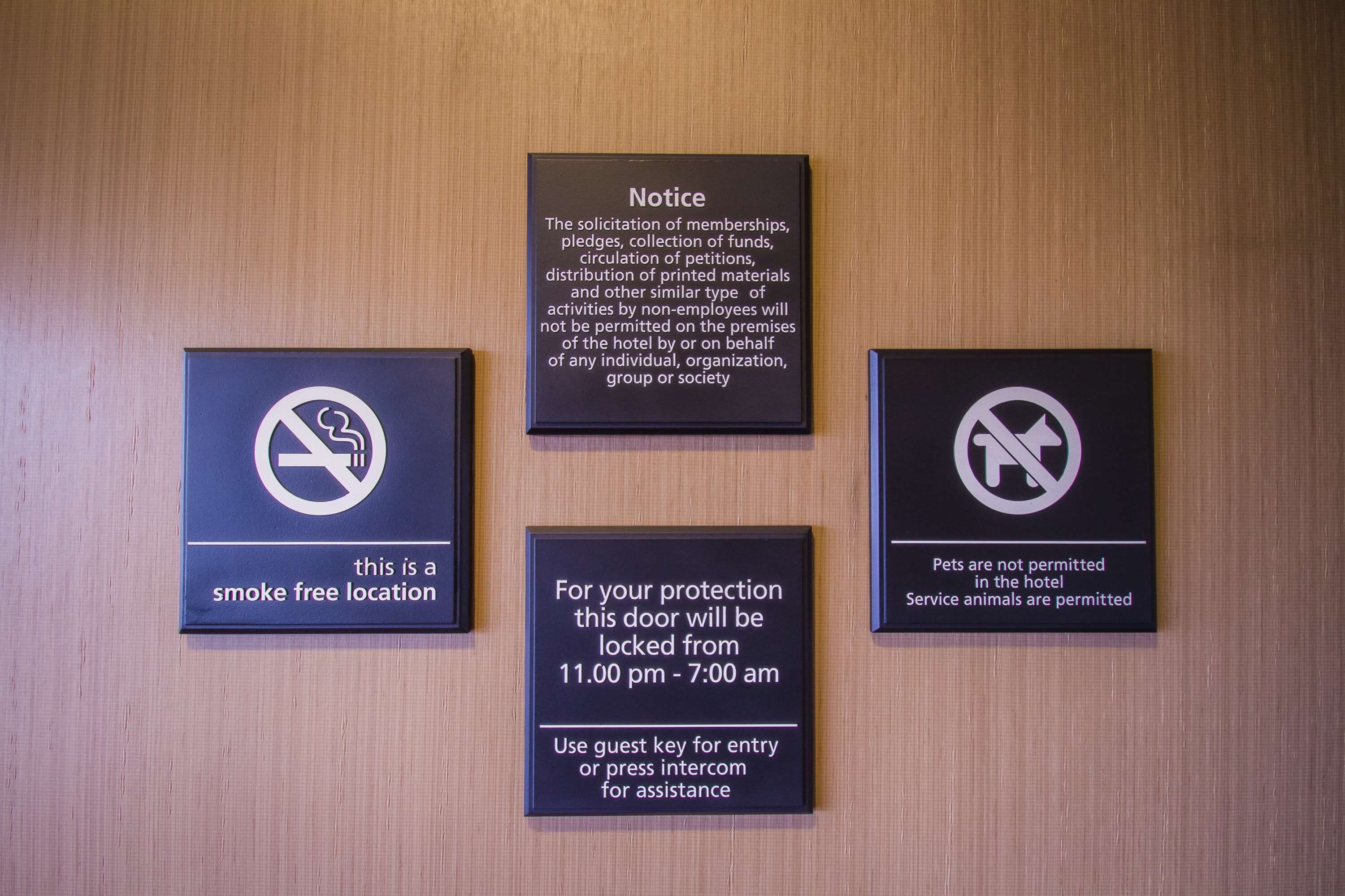 Four signs on a wooden wall: a smoke-free location, a notice about non-employee activities, a locked door from 11:00 pm to 7:00 am, and a pet policy allowing service animals.