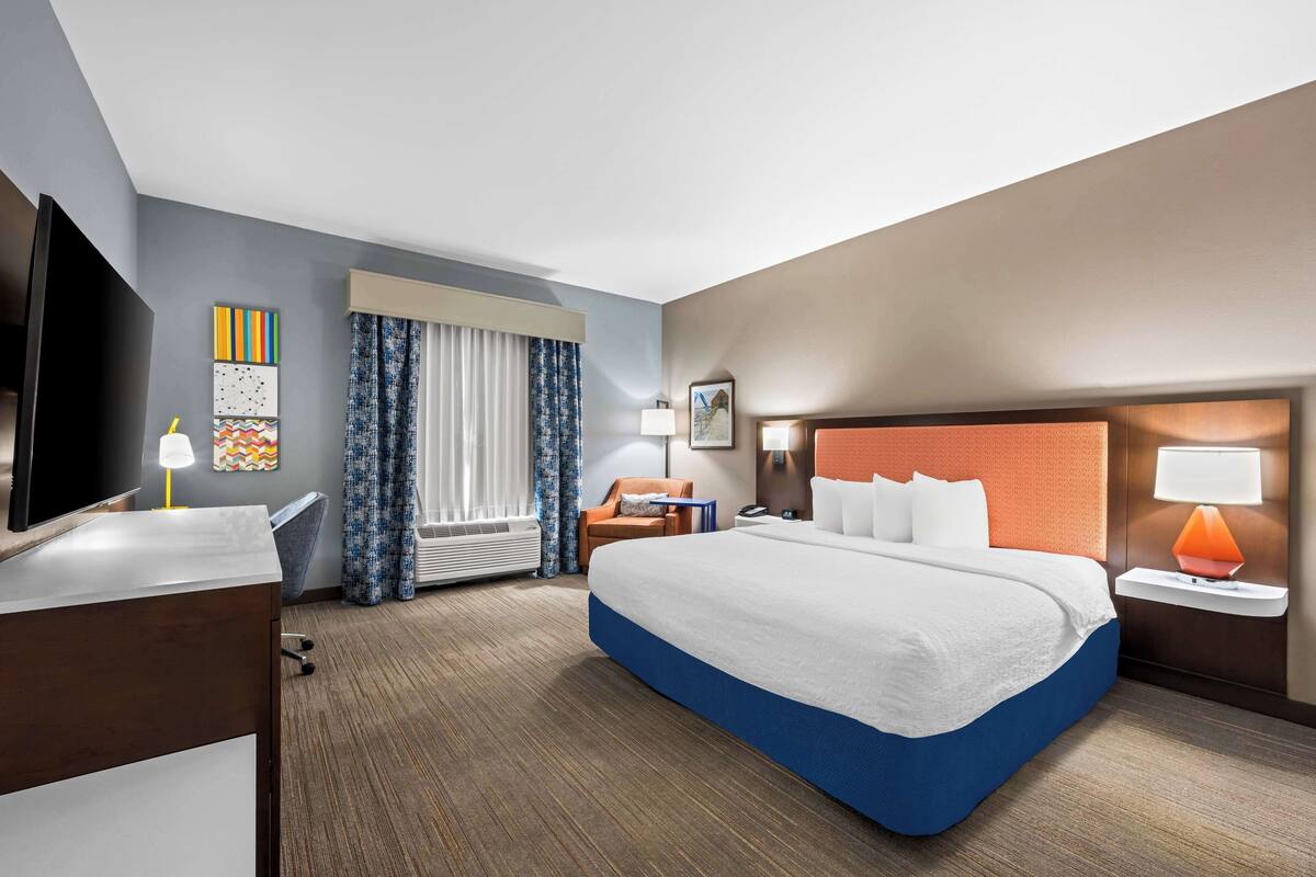 A hotel room with a large bed, a desk with a lamp, a chair, a TV, and a window with curtains.