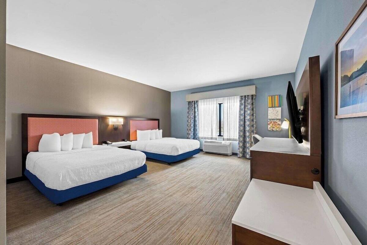 A hotel room with two beds, a desk, a chair, a window with curtains, and a painting on the wall.