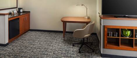 A hotel room with a kitchenette, a desk with a chair, a television, and a small cabinet with glassware.