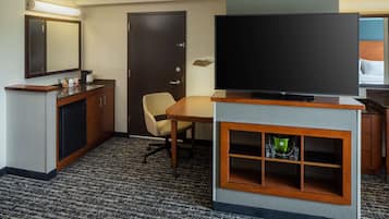 A hotel room with a flat-screen TV, a desk, a chair, a minibar, and a door.