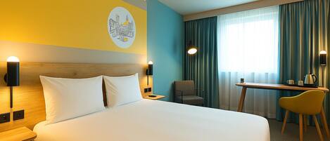 Habitación de hotel con una cama grande, un escritorio, dos sillas, una lámpara de pared y un ventanal con cortinas.
