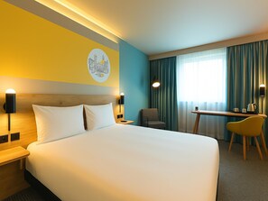 Une chambre d’hôtel avec un grand lit, un bureau, deux chaises, une lampe fixée au mur et une fenêtre avec des rideaux.