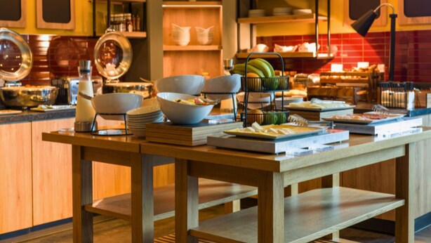 Un buffet est installé sur une table en bois, avec différents plats et saladiers disposés dessus, et entouré d’étagères remplies d’ustensiles de cuisine.