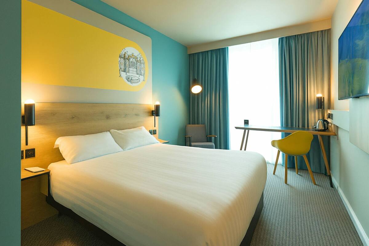 A hotel room with a large bed, a desk with a chair, a painting on the wall, and a window with curtains.