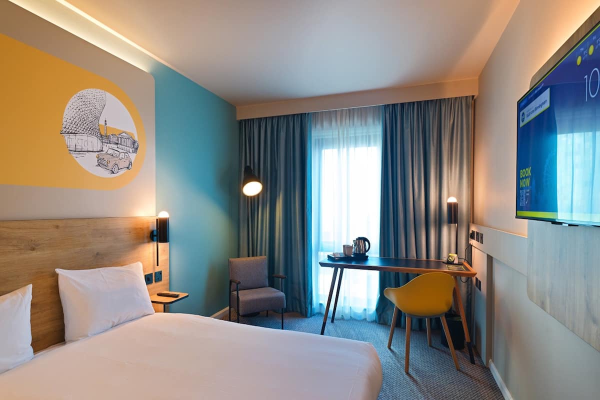 A hotel room with a large bed, a desk, a chair, a TV, and a window with curtains.