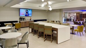A modern hotel lobby with a central bar area, seating arrangements, and a television screen displaying a show. - La Quinta Inn & Suites by Wyndham Port Arthur (Port Arthur)