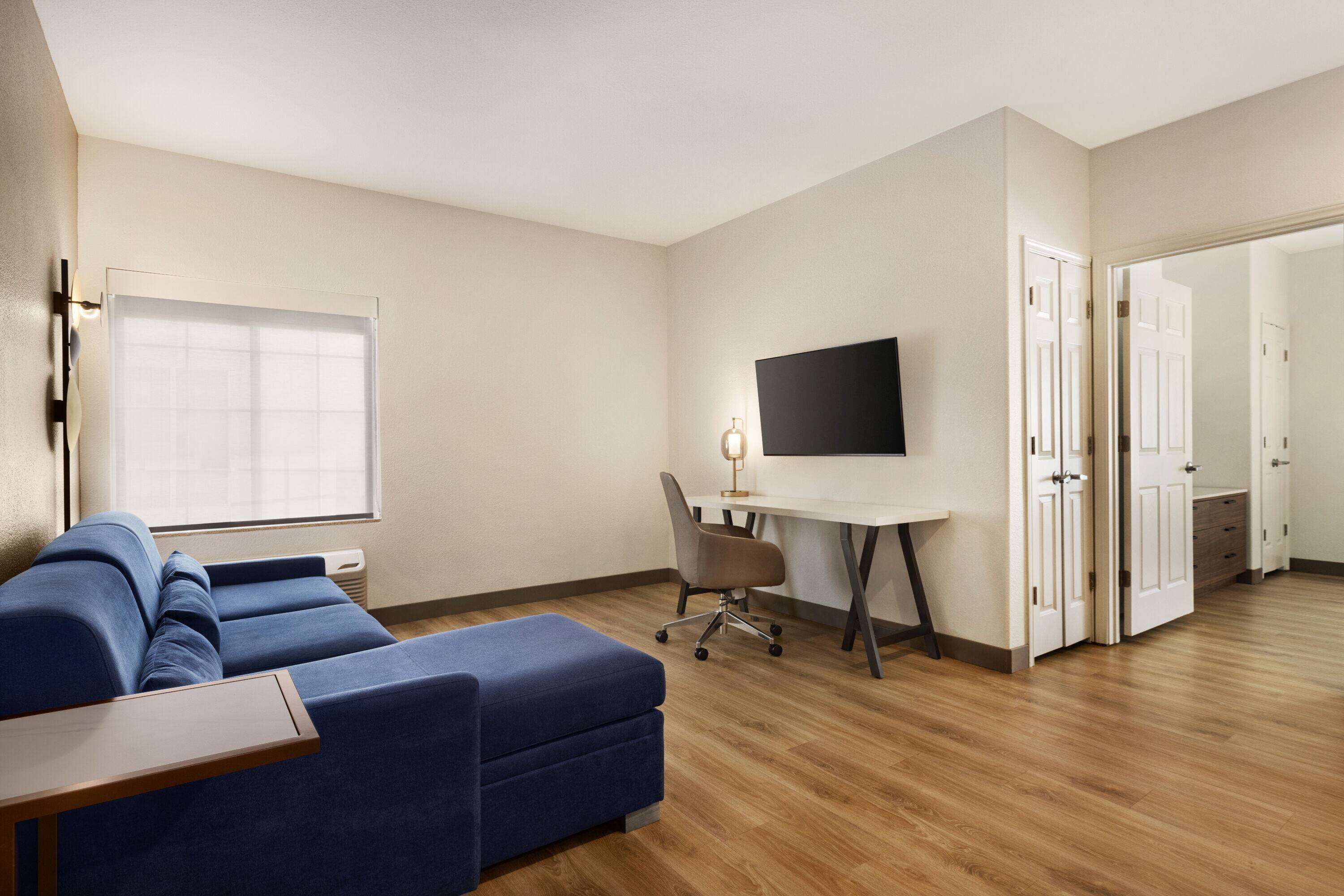 A modern living room with a blue sofa, a wooden coffee table, a desk with a chair, a flat-screen TV, and a large window.