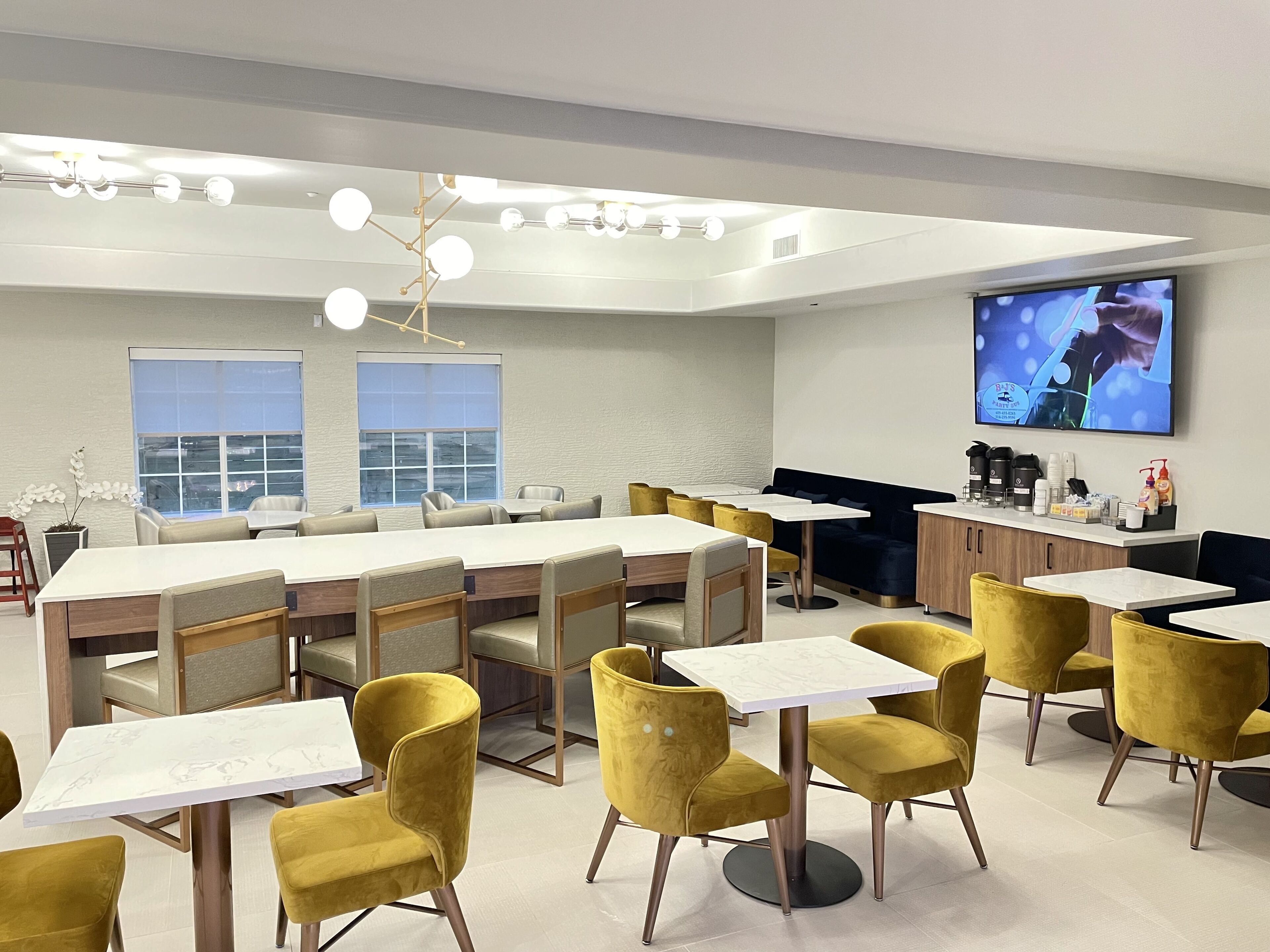 A modern dining area with yellow chairs and white tables, a bar area with a TV, and a large window providing natural light.